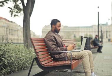 Bilgisayarı ve akıllı telefonu olan bir bankta oturan genç adam.