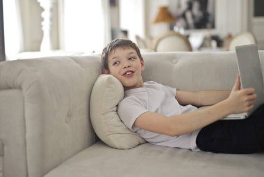 Elinde bilgisayar olan bir çocuk kanepede yatıyor.