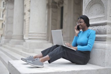 Okulun merdivenlerindeki genç kadın bilgisayar ve akıllı telefonla çalışıyor.