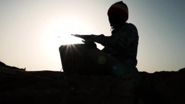 Afrika perküsyon müziği, Senegalli bir adamın silueti günbatımında elleriyle geleneksel bir djembe davul çalıyor. Fas, Essaouira plajında açık hava müziği. 4k