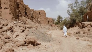 Geleneksel giyinmiş Faslı bir adam beyaz gandoura, djellaba ve türban giyerek Tamnougalt, Fas 'ta bir Kasbah' ın önünde yürüyor. Güney Fas 'ın otantik kırsal manzarası. 4k.