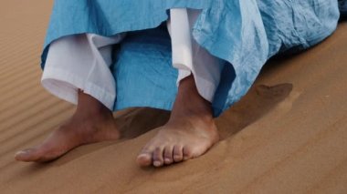 A traditionally dressed Moroccan man wearing a blue gandoura, djellaba sits barefoot on a sand dune, resting, waiting. Authentic desert scene of South Morocco. 4k.