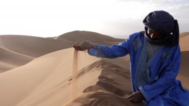 A Moroccan man, wearing a blue gandoura, djellaba and turban, rests on top of a sand dune, Chigaga desert, Morocco. Authentic Bedouin of South Morocco. 4k.