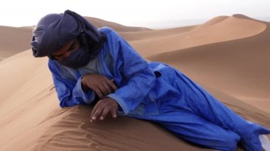 A Moroccan man, wearing a blue gandoura, djellaba and turban, rests on top of a sand dune, Chigaga desert, Morocco. Authentic Bedouin of South Morocco. 4k.