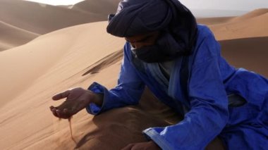 Mavi gandoura, djellaba ve türban takan Faslı bir adam kumla oynayan bir kum tepesinde, Chigaga çölünde, Fas 'ta yatıyor. Güney Fas 'ın otantik bedevileri. 4k görüntü.