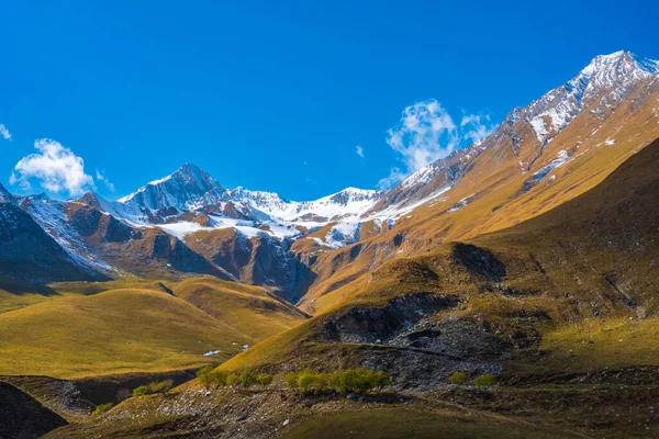 Soluk kesici dağ zirvelerinin alçak açısı ve parlak güneş ışığı altında küçük bulutlu mavi gökyüzüne bakan yeşil çimenli tepeler.