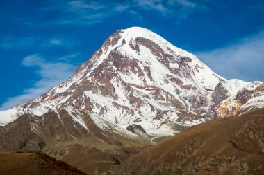 Karla kaplı Kazbegi Dağı 'nın nefes kesici manzarası kırsalda beyaz bulutlu mavi gökyüzüne karşı.