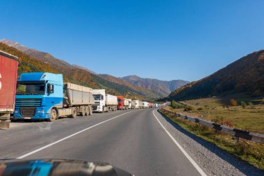 Güneşli bir günde, dağ sırtı ve bulutsuz mavi gökyüzüne karşı asfalt yol kenarında sıraya girmiş bir sürü kamyon var.