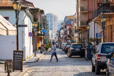 Batumi, Gürcistan - 12 Mayıs 2022: Güneşli bir günde Gürcistan 'ın başkenti Batumi' de evleri olan eski şehir sokağındaki modern arabaların yakınındaki kaldırım taşı yoldan geçen adam