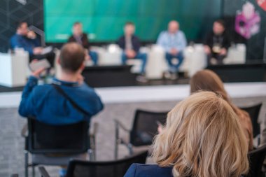 Sarı saçlı, meslektaşları ve gazetecilerle birlikte sandalyede oturan ve iş semineri sırasında sahnede hoparlörleri dinleyen tanınmamış kadın yöneticinin arka planı.