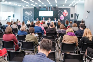 Çağdaş konferans salonundaki seminer sırasında tanımadığımız kadın ve erkeklerin resmi kıyafetlerle sandalyelere oturup alet kullanmaları ve hoparlörleri dinlemeleri.