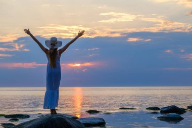 Beyaz elbiseli anonim bir kadın deniz kenarında duruyor ve gün batımında kollarını açıp özgürlüğün tadını çıkarıyor.