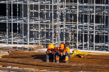 Metal çubukların üzerinde oturan ve inşaat halindeyken cep telefonlarını karıştıran koruyucu üniformalı tam vücut adamları.