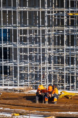 Metal çubukların üzerinde oturan ve inşaat halindeyken cep telefonlarını karıştıran koruyucu üniformalı tam vücut adamları.