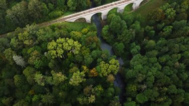 Yukarıdan, kırsal bölgedeki yeşil ormanlık alanda demiryolu köprüsünün altından akan dar nehir manzarası.