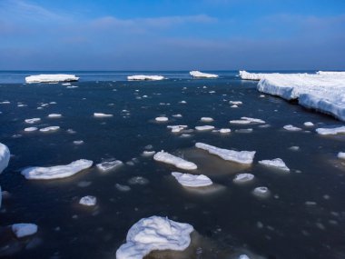 Karanlık deniz suyunda eriyen buz kütleleri, dondurulmuş kıyılarla güneşli havada bulutlu mavi gökyüzünün altında yüzüyor.