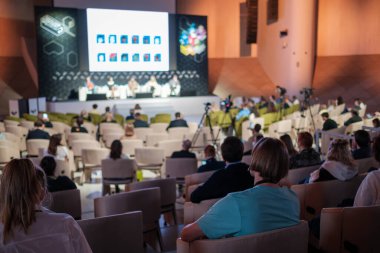 Modern salondaki iş semineri sırasında sandalyelerde oturan ve hoparlörleri dinleyen isimsiz insanların arka plan görüntüsü.