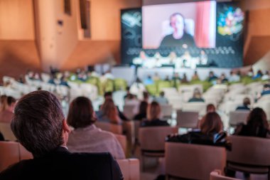 Sandalyelerde oturan ve geniş oditoryumdaki iş konferansı sırasında büyük ekrandan konuşan kadınları dinleyen isimsiz insanlar.