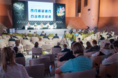 Modern salondaki iş semineri sırasında sandalyelerde oturan ve hoparlörleri dinleyen isimsiz insanların arka plan görüntüsü.