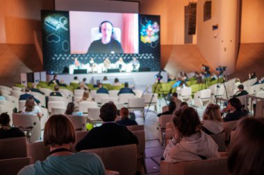 Sandalyelerde oturan ve geniş oditoryumdaki iş konferansı sırasında büyük ekrandan konuşan kadınları dinleyen isimsiz insanlar.