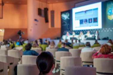 Modern salondaki iş semineri sırasında sandalyelerde oturan ve hoparlörleri dinleyen isimsiz insanların arka plan görüntüsü.
