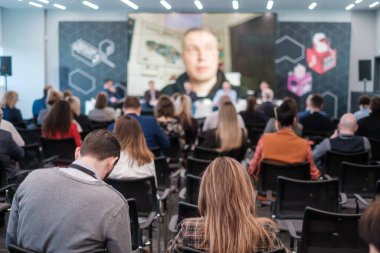 Geniş oditoryumdaki iş konferansı sırasında sandalyelerde oturan ve büyük ekrandan erkek hoparlörleri dinleyen isimsiz insanlar