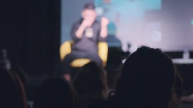 Karanlık oditoryumdaki konferans sırasında sahne arkasından konuşan isimsiz insanların siluetleri.