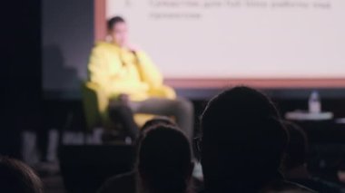 Karanlık oditoryumdaki konferans sırasında sahne arkasından konuşan isimsiz insanların siluetleri.