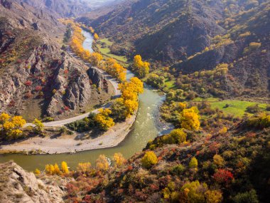 Güneşli bir günde, dağlık arazide, hızlı nehir ve sonbahar ağaçlarının yakınındaki virajlı asfalt yolda giden karavanın insansız hava aracı görüntüsünden.
