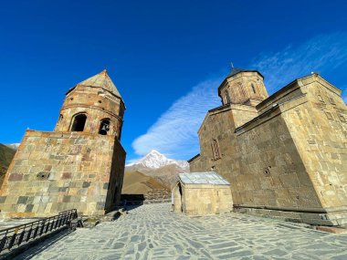Gürcistan 'ın dağlık kesimlerinde güneşli bir günde bulutlu mavi gökyüzüne karşı kurulan ortaçağ Gergeti Trinity Kilisesi manzarası