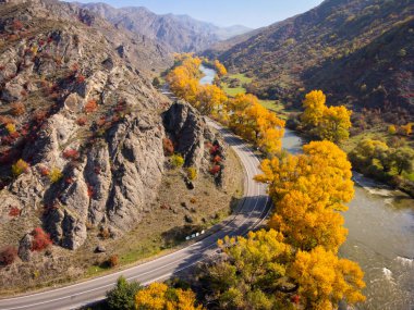 Güneşli bir günde, dağlık arazide, hızlı nehir ve sonbahar ağaçlarının yanındaki kavisli asfalt yolun yukarıdan insansız hava aracı görüntüsünden.