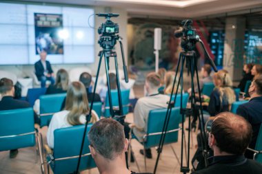 Profesyonel kameraların yanındaki mavi koltuklarda oturan isimsiz insanlar tripodlu ve modern konferans salonunda sunum sırasında hoparlör dinliyorlar.