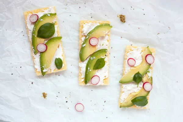 Beyaz arka planda yumuşak peynir, avokado, turp ve fesleğen ile glütensiz kızarmış ekmek.