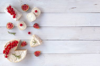 Camembert ve keçi peyniri beyaz ahşap arka planda incir, kırmızı frenk üzümü ve biberiye dilimleri ile süslenmiş, üst manzara, kopyalama alanı