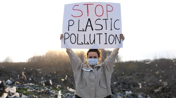 Sad woman volunteer activist stands at big landfill site with garbage with Stop Plastic Pollution poster demonstrates against protection environment. Pollution and ecology.