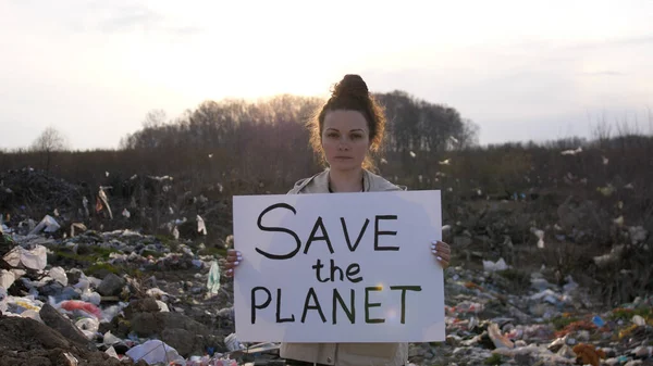 Sad woman volunteer activist stands at big landfill site with garbage with Save the Planet poster demonstrates against protection environment. Pollution and ecology.