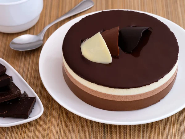 A Chocolate mousse cake on a white plate. Plate with pieces of chocolate, teaspoon and cup near it on a bamboo napkin