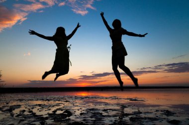 Gün batımında heyecanla atlayan iki genç kadının silueti.