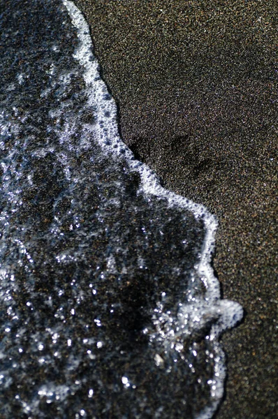 Human footprint on black sand. Black volcanic magnetic sand on the seashore. Background texture of the beach with waves closeup.