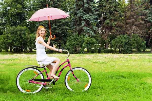 Young woman riding a bicycle in a park holding a pink umbrella — Stock Photo, Image