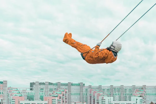 Astronauta usando terno de espaço laranja e capacete espacial voando em um balanço contra o céu nublado acima da paisagem urbana — Fotografia de Stock