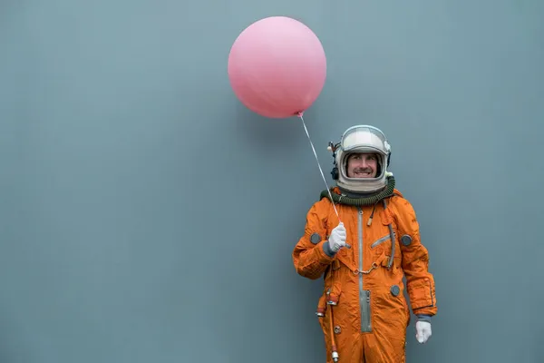 Astronaut in Raumanzug und Helm, der einen rosafarbenen aufblasbaren Ballon vor grauen Wandhintergrund hält. Glücklicher Kosmonaut mit Helium-Luftballon — Stockfoto