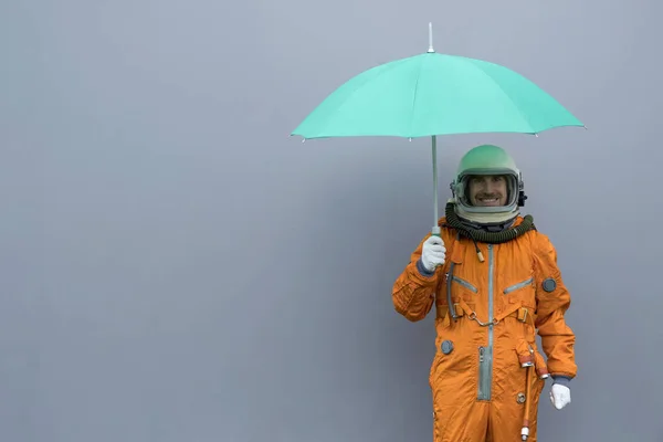 Glücklicher Astronaut in orangefarbenem Raumanzug und Weltraumhelm, der einen offenen grünen Regenschirm vor grauem Wandhintergrund hält. Versicherungskonzept — Stockfoto