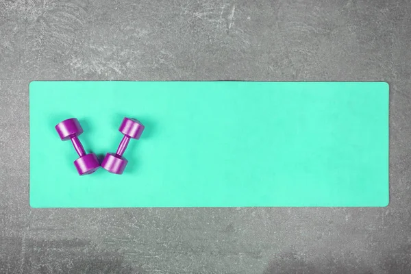 Плоский слой бирюзового коврика для йоги с фиолетовыми гантелями на бетонном полу. Пара гантели на фитнес-коврик разбросаны по цементному полу — стоковое фото