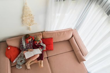 Anne ve kızı pijama giyer ve geyik boynuzu takar. Yılbaşı bandı takar ve kitap okurlar. Yukarıdan bak. Kanepede otururken küçük kızını kucaklayan genç bir kadın. Üst görünüm