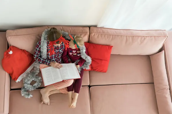 Madre con hija vistiendo pijamas y cuernos de cornamenta Diademas de Navidad mientras lee un libro. Mujer joven abrazando a su niña mientras se relaja en el sofá y leyendo un libro — Foto de Stock