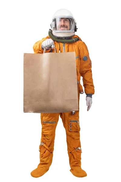 Happy astronaut wearing orange space suit and space helmet holding in hand blank kraft paper bag isolated on white background. Delivery concept — Stock Photo, Image
