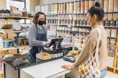 Çevre çantalı Afrikalı Amerikalı bir kadın kredi kartıyla ödeme yapıyor. Sürdürülebilir yerel bir dükkanda, tezgahın önünde duruyor. Ellere ve POS terminaline odaklan
