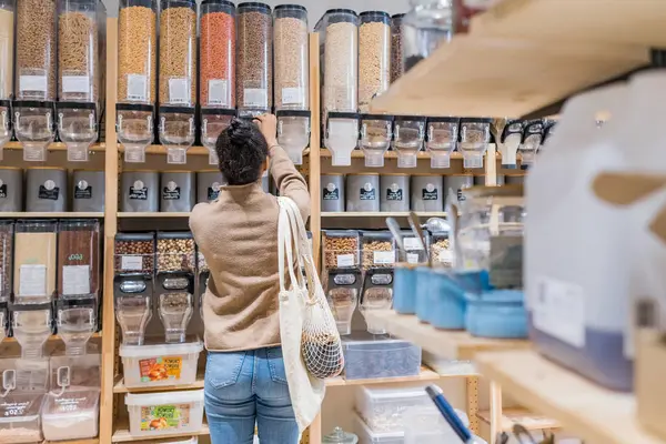 Donna afroamericana che compra cereali e cereali biologici in un supermercato sostenibile a zero rifiuti. Giovane donna che riempie il contenitore riutilizzabile nel negozio di alimentari locale — Foto Stock