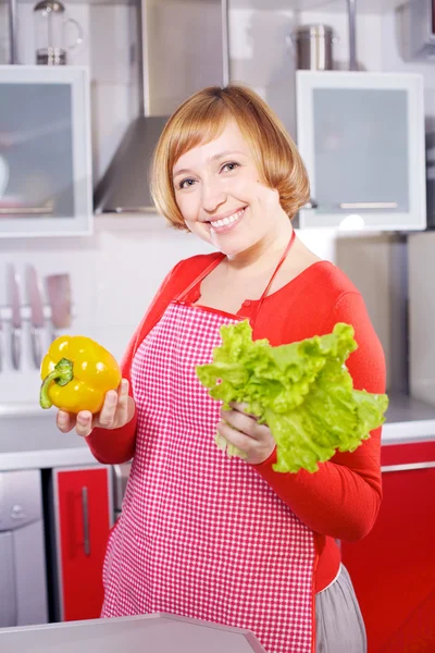 Jovem dona de casa bonita na cozinha vermelha segurando pimenta e alface — Fotografia de Stock